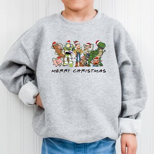 May include: A gray sweatshirt with a Christmas-themed design featuring characters from the Toy Story franchise. The design includes Woody, Buzz Lightyear, Jessie, Rex, and other characters. The text "Merry Christmas" is printed below the characters.