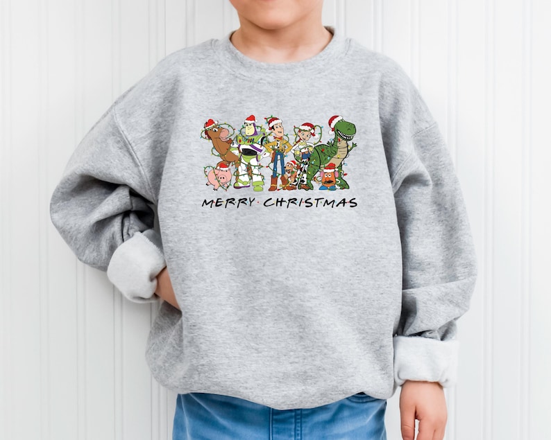 May include: A gray sweatshirt with a Christmas-themed design featuring characters from the Toy Story franchise. The design includes Woody, Buzz Lightyear, Jessie, Rex, and other characters. The text "Merry Christmas" is printed below the characters.