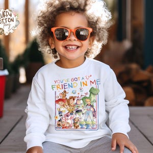 Op de afbeelding: Een witte longsleeve trui met een kleurrijke afbeelding van "Toy Story"-personages en de tekst "YOU'VE GOT A FRIEND IN ME". Het kind draagt een oranje zonnebril. Geschikt voor kinderen.