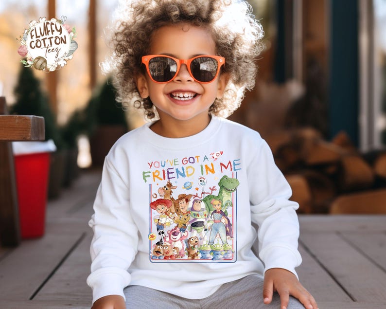 Op de afbeelding: Een witte longsleeve trui met een kleurrijke afbeelding van "Toy Story"-personages en de tekst "YOU'VE GOT A FRIEND IN ME". Het kind draagt een oranje zonnebril. Geschikt voor kinderen.