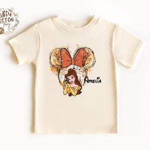 以下が含まれることがあります： クリーム色のTシャツ。美女と野獣のベルの漫画イラストが描かれています。デザインにはリボン、花のアクセント、そして「Amelia」という名前が黒い筆記体で書かれています。Tシャツは半袖です。
