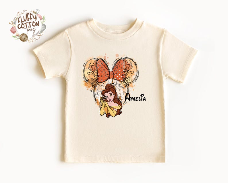 以下が含まれることがあります： クリーム色のTシャツ。美女と野獣のベルの漫画イラストが描かれています。デザインにはリボン、花のアクセント、そして「Amelia」という名前が黒い筆記体で書かれています。Tシャツは半袖です。