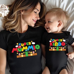 May include: Black t-shirts with colorful text and graphics. One shirt says "SUPER MOMMIO" and the other says "SUPER KIDDO". The graphics are inspired by a video game theme, with clouds, mushrooms, and a green pipe.
