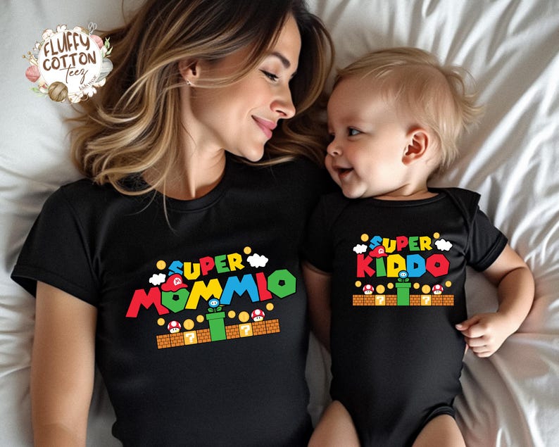 May include: Black t-shirts with colorful text and graphics. One shirt says "SUPER MOMMIO" and the other says "SUPER KIDDO". The graphics are inspired by a video game theme, with clouds, mushrooms, and a green pipe.
