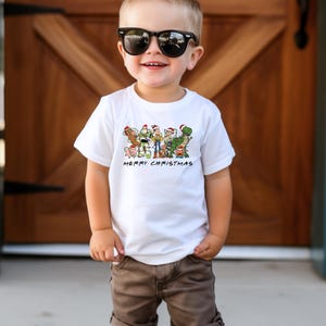 May include: A white t-shirt with a graphic of the Toy Story characters wearing Santa hats. The text "Merry Christmas" is printed below the graphic. A young child is wearing the shirt and has brown hair and is wearing sunglasses.