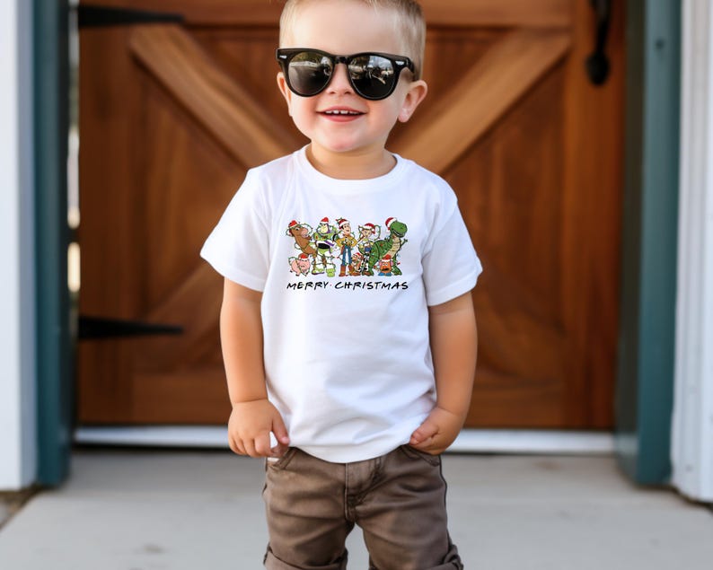 May include: A white t-shirt with a graphic of the Toy Story characters wearing Santa hats. The text "Merry Christmas" is printed below the graphic. A young child is wearing the shirt and has brown hair and is wearing sunglasses.