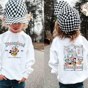 May include: A white sweatshirt with "Disneyland" in teal script and a graphic of Disney characters. The front of the shirt has the text "EST 1955 CALIFORNIA". The child is wearing a black and white checkered baseball cap.