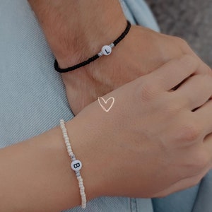Könnte beinhalten: Zwei Freundschaftsarmbänder mit weißen Perlen und schwarzem Band. Ein Armband hat eine weiße Perle mit dem Buchstaben "L" darauf, das andere eine weiße Perle mit dem Buchstaben "B" darauf.