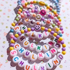 May include: A collection of colourful beaded bracelets. Each bracelet features white letter beads spelling "SCHULKIND" and smiley face beads. The beads are in various colours, including blue, pink, yellow, and purple. The bracelets are stacked on a light background.