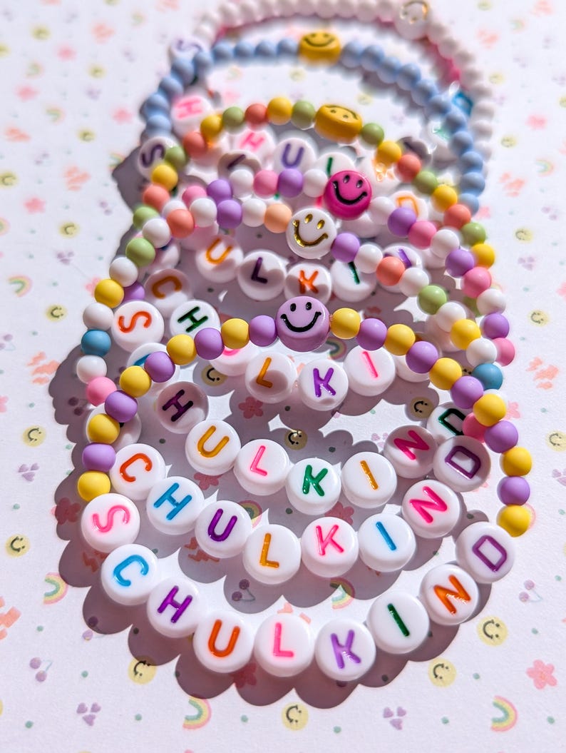May include: A collection of colourful beaded bracelets. Each bracelet features white letter beads spelling "SCHULKIND" and smiley face beads. The beads are in various colours, including blue, pink, yellow, and purple. The bracelets are stacked on a light background.