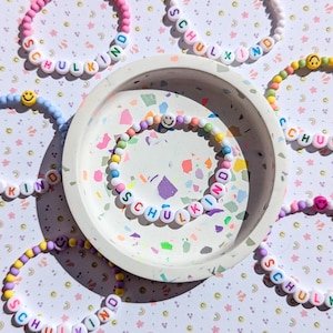 May include: A selection of colourful beaded bracelets, each spelling out "SCHULKIND" or "KIND" with letter beads. Smiley face beads are incorporated. The bracelets are displayed on a decorative tray, with a patterned background.