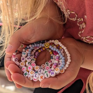 May include: A selection of colourful beaded bracelets spelling out "SCHOOL" and "KIND" in white letter beads. The bracelets are held in a person's hands, with a pink top and blonde hair in the background. Smiley face beads are also included.