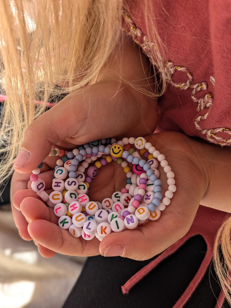 May include: A selection of colourful beaded bracelets spelling out "SCHOOL" and "KIND" in white letter beads. The bracelets are held in a person's hands, with a pink top and blonde hair in the background. Smiley face beads are also included.