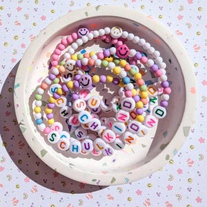 May include: A selection of colourful beaded bracelets with smiley face beads and letter beads spelling "SCHULKIND" in a speckled tray. The bracelets are in shades of pink, yellow, purple, and blue.