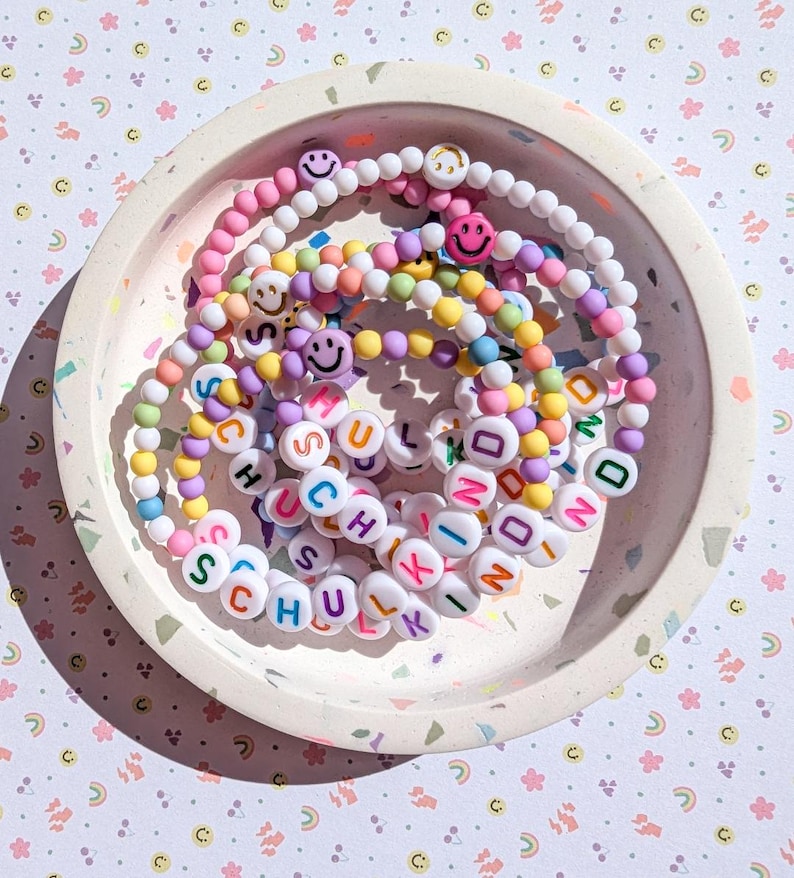 May include: A selection of colourful beaded bracelets with smiley face beads and letter beads spelling "SCHULKIND" in a speckled tray. The bracelets are in shades of pink, yellow, purple, and blue.