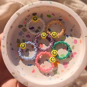 Könnte beinhalten: Ein Set aus sechs bunten Perlenringen mit einem gelben Smiley-Anhänger an jedem Ring. Die Ringe werden in einer weißen, gefleckten Schale präsentiert.