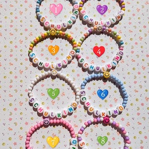 May include: Eight colourful beaded bracelets spelling "SCHULKIND" with white letter beads. Each bracelet has a different colour scheme, including blue, purple, yellow, and pink, and smiley face beads.