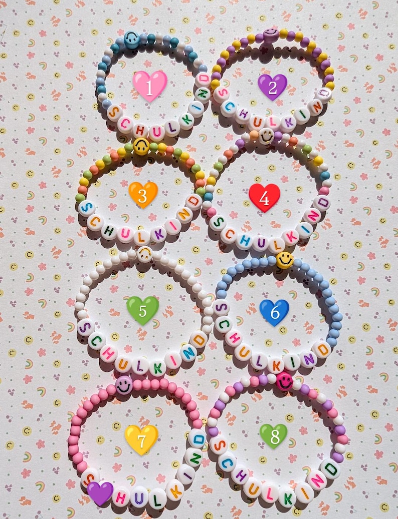 May include: Eight colourful beaded bracelets spelling "SCHULKIND" with white letter beads. Each bracelet has a different colour scheme, including blue, purple, yellow, and pink, and smiley face beads.