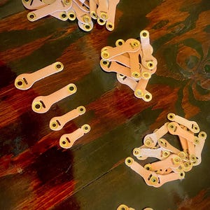 May include: Several small, light brown leather pieces with brass eyelets are scattered on a dark wooden surface. The pieces are connected in a chain-like fashion, suggesting a crafting or DIY project. The eyelets provide a point of connection.