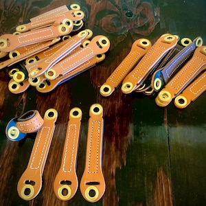 May include: A collection of tan leather straps with gold-colored eyelets and white stitching. The straps are arranged on a dark wooden surface. Some straps have a blue interior.