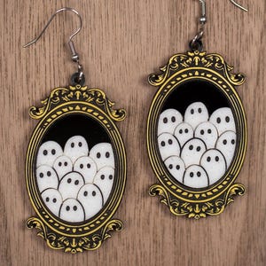Op de afbeelding: Een paar Halloween-oorbellen. Elke oorbel heeft een ovale lijst met een zwarte achtergrond en een cluster van witte spookgezichten. De lijsten zijn goudkleurig omlijnd en hebben decoratieve details, met zilverkleurige oorhaken.