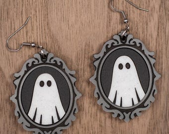 May include: A pair of Halloween-themed earrings. Each earring features a white ghost design within a black oval frame, set against a gray decorative border. The earrings hang from silver-colored ear wires, perfect for spooky season.