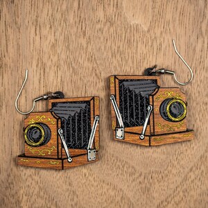 May include: Wooden earrings shaped like vintage cameras. The earrings feature a detailed design with black bellows, gold accents, and silver-colored hooks. The earrings are set against a wooden background.
