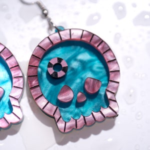May include: A pair of skull-shaped earrings. The earrings feature a teal interior with a pink and brown border. The eyes and mouth are also pink and brown. The earrings are hanging from silver hooks.