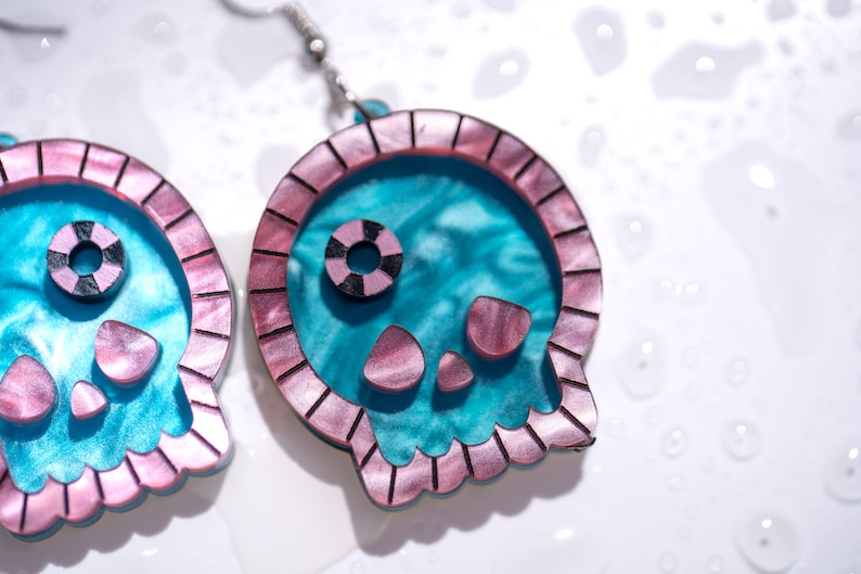 May include: A pair of skull-shaped earrings. The earrings feature a teal interior with a pink and brown border. The eyes and mouth are also pink and brown. The earrings are hanging from silver hooks.