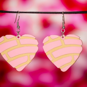 May include: A pair of pink and gold heart-shaped earrings. The earrings are shaped like a traditional Mexican sweet bread called a concha.