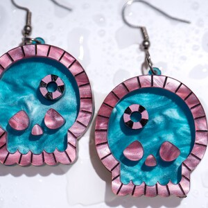 May include: A pair of skull-shaped earrings. The earrings feature a teal and pink color scheme. The skull's outline is pink, with a teal interior. The eyes are black and pink. The earrings are suspended from silver-colored hooks.