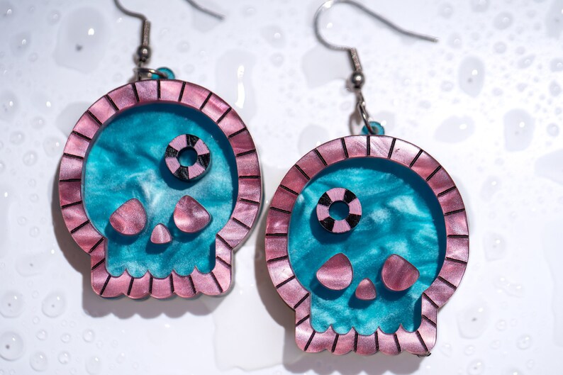 May include: A pair of skull-shaped earrings. The earrings feature a teal and pink color scheme. The skull's outline is pink, with a teal interior. The eyes are black and pink. The earrings are suspended from silver-colored hooks.