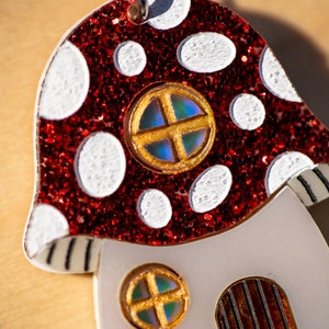 May include: A red and white mushroom-shaped ornament with glitter and a gold-trimmed window. The ornament has a white base with two additional windows.