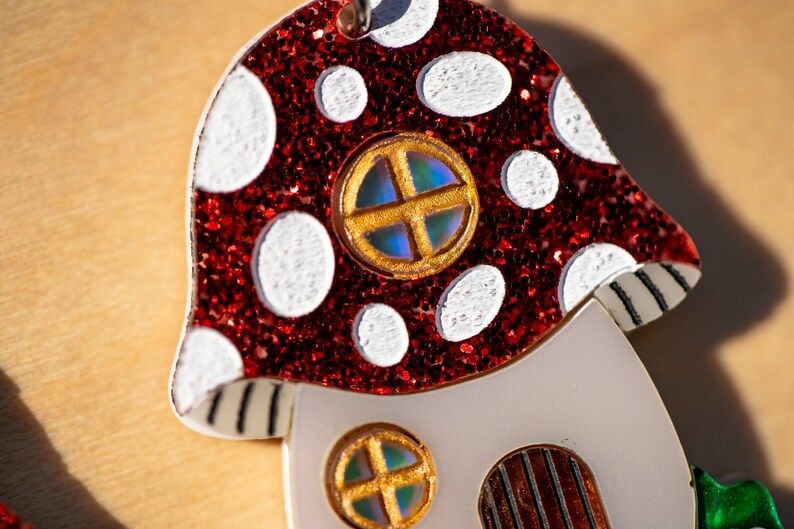 May include: A red and white mushroom-shaped ornament with glitter and a gold-trimmed window. The ornament has a white base with two additional windows.