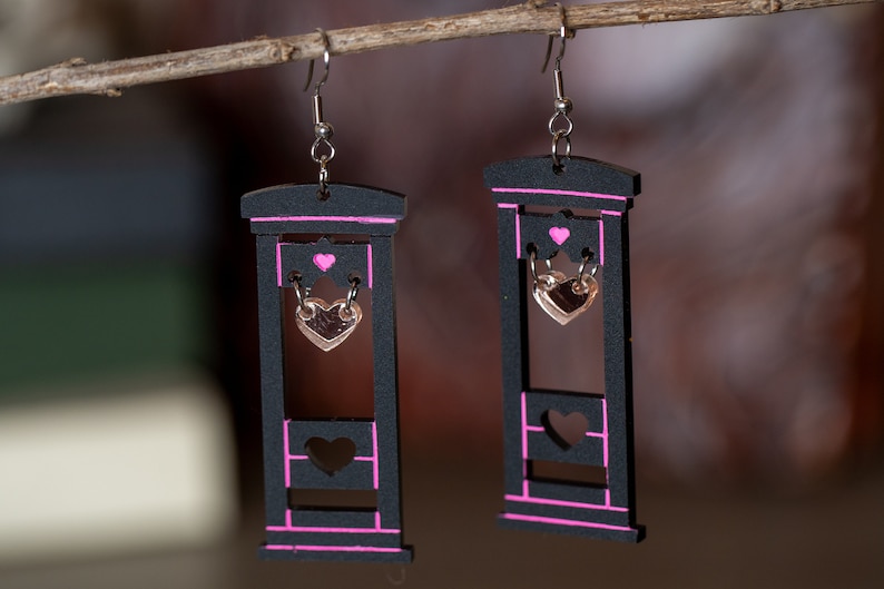 May include: A pair of black wooden earrings shaped like a guillotine with pink accents. Each earring has a small gold heart hanging from the blade.