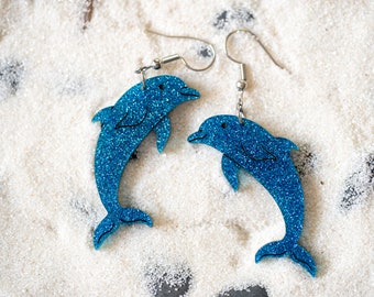 Könnte beinhalten: Ein Paar blaue Glitzer-Delfin-Ohrringe. Die Delfine sind in entgegengesetzte Richtungen gerichtet und an silbernen Haken aufgehängt.