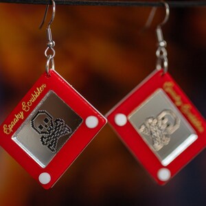 May include: A pair of red earrings shaped like a red Etch A Sketch toy with a silver skull and crossbones drawing on the screen. The earrings are labeled "Spooky Scribbler" and "Creepy Crawler".