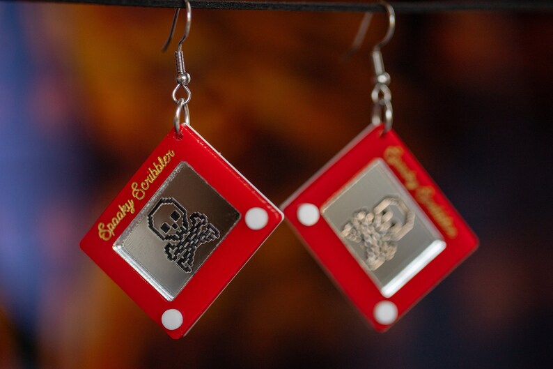 May include: A pair of red earrings shaped like a red Etch A Sketch toy with a silver skull and crossbones drawing on the screen. The earrings are labeled "Spooky Scribbler" and "Creepy Crawler".