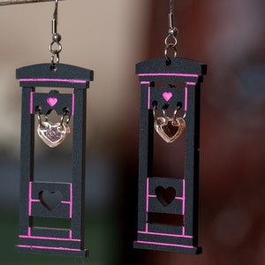May include: A pair of black wooden earrings shaped like a guillotine with pink accents. Each earring features a dangling heart charm.