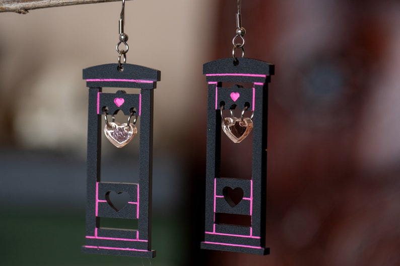 May include: A pair of black wooden earrings shaped like a guillotine with pink accents. Each earring features a dangling heart charm.