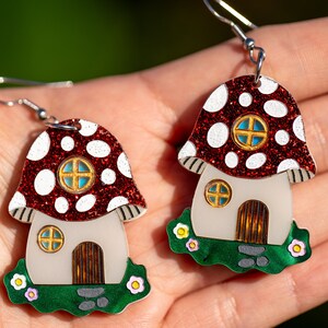 May include: A pair of red and white mushroom house earrings with glitter accents. The houses have white walls, green grass, and brown doors.