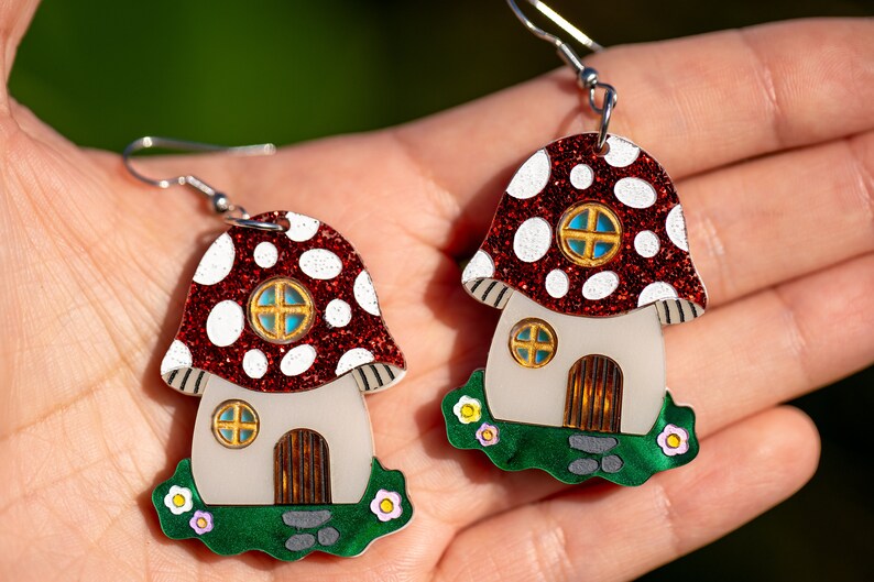 May include: A pair of red and white mushroom house earrings with glitter accents. The houses have white walls, green grass, and brown doors.