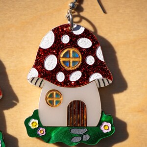 May include: A pair of red and white mushroom-shaped earrings with a white house and green grass. The house has a brown door and two windows with gold frames.