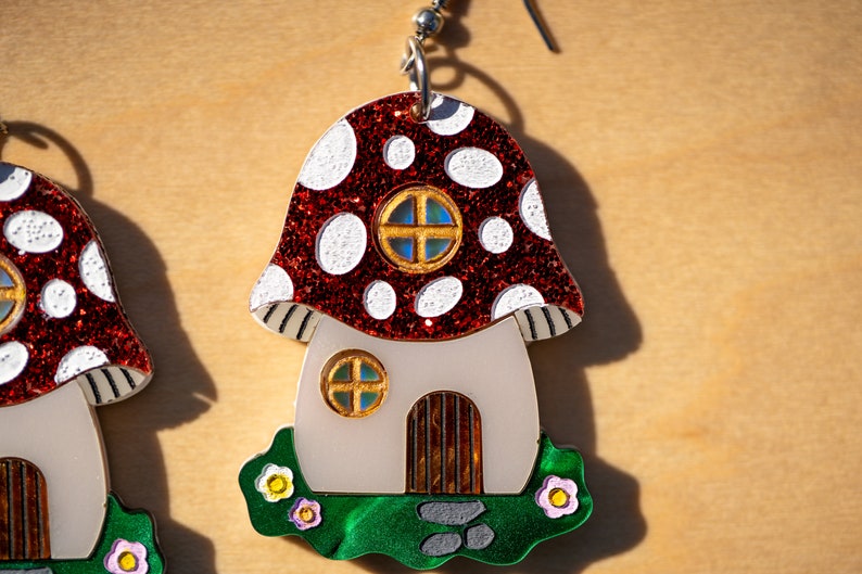 May include: A pair of red and white mushroom-shaped earrings with a white house and green grass. The house has a brown door and two windows with gold frames.