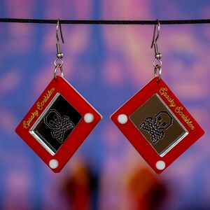May include: A pair of red earrings shaped like a vintage Etch A Sketch toy with a skull and crossbones design and the text "Spooky Scribbler" on the side.
