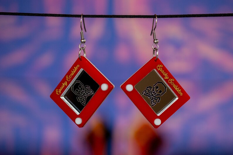 May include: A pair of red earrings shaped like a vintage Etch A Sketch toy with a skull and crossbones design and the text "Spooky Scribbler" on the side.