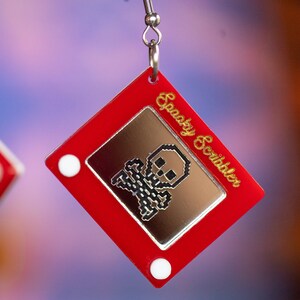 May include: Red plastic earrings shaped like a vintage Etch A Sketch toy with a pixelated skull and crossbones image and the text "Spooky Scribbler" on the side.