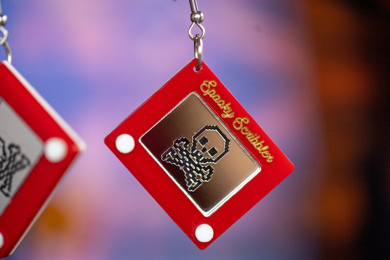 May include: Red plastic earrings shaped like a vintage Etch A Sketch toy with a pixelated skull and crossbones image and the text "Spooky Scribbler" on the side.