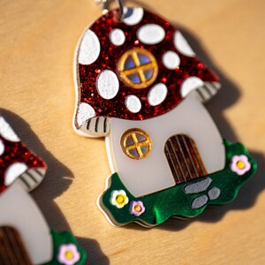 May include: A pair of red and white mushroom-shaped earrings with a white house and green grass base. Each house has a brown door and a window with a gold frame and iridescent glass.