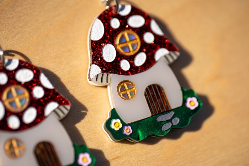 May include: A pair of red and white mushroom-shaped earrings with a white house and green grass base. Each house has a brown door and a window with a gold frame and iridescent glass.